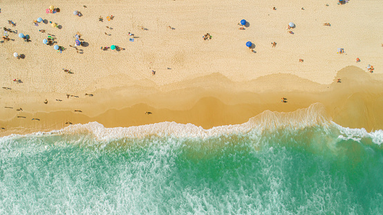 Aerial locked shot of waves breaking on the shore. Colourful beach umbrellas and people enjoying the summer.
