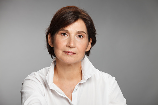 Middle aged woman portrait wearing white shirt against gray solid background.