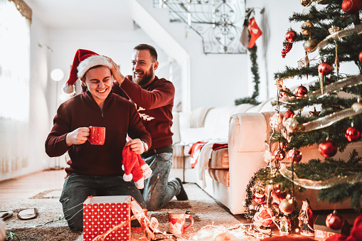 Smiling Gay Couple Opening Christmas Presents With Santa Hats And Drinking Tea Together