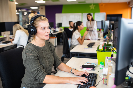 A young woman is talking trough a headset while working on her computer at the office