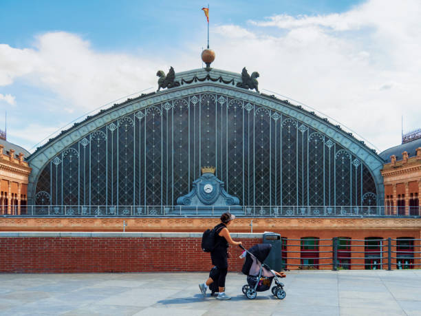 Madrid, Spain. 02/09/2021 A woman with her child passing in front of the main facade of Madrid's Atocha station on a cloudy sky day.