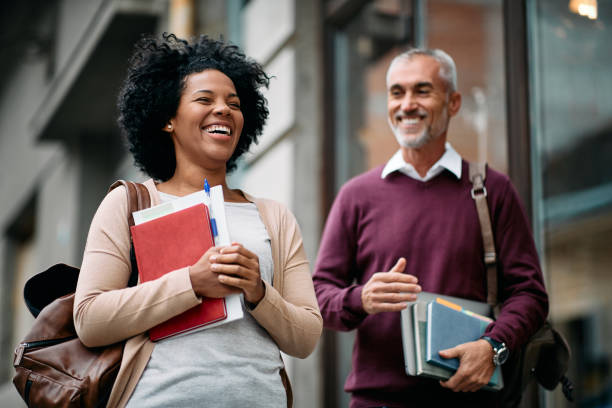Happy adult students laughing and having fun in front of university library. Happy mid adult African American students and her mature friend having fun while going to a library to study. adult education stock pictures, royalty-free photos & images