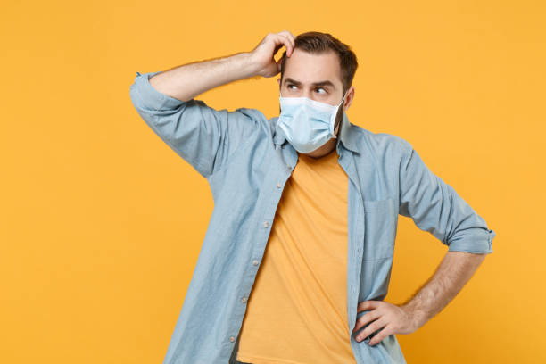 Puzzled young man in sterile face mask posing isolated on yellow background. Epidemic pandemic rapidly spreading coronavirus 2019-ncov sars covid-19 flu virus concept. Looking aside, put hand on head joven desconcertado con máscara facial estéril posando aislado sobre fondo amarillo. pandemia epidémica que se propaga rápidamente por el coronavirus 2019-ncov sars covid-19 concepto del virus de la gripe. mirando a un lado, pon la mano en la cabeza.