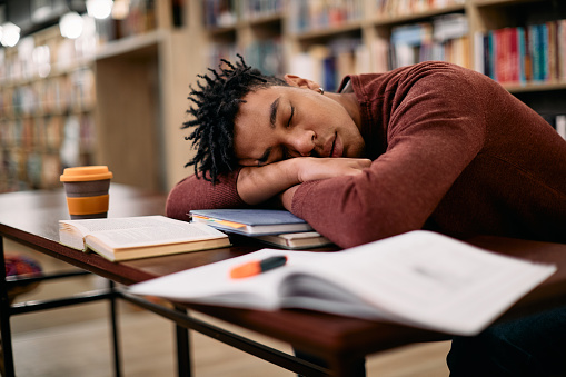 Exhausted African American student taking a nap while studying in a library.