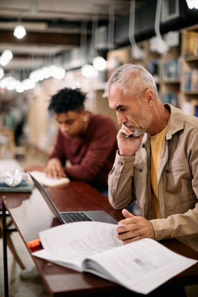 Mature male student using laptop while studying in a library. Mature man using laptop while studying and doing a research in a library. middle age adult studying stock pictures, royalty-free photos & images