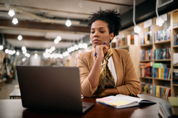 Mid adult black university professor working on laptop in a library. African American female professor using laptop while doing a research at university library. lecturer stock pictures, royalty-free photos & images