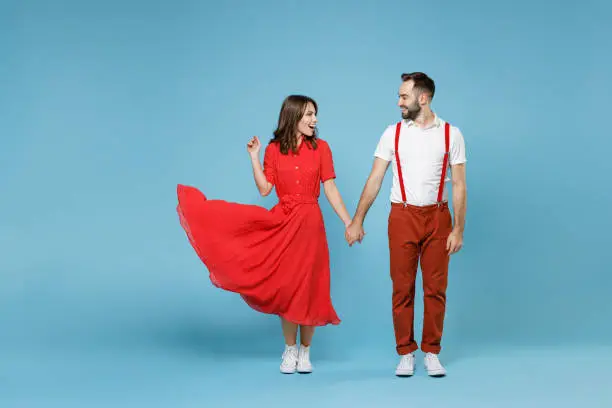 Full length smiling young couple two friends man woman in white red clothes holding hands looking at each other isolated on pastel blue background studio portrait. St. Valentine's Day holiday concept