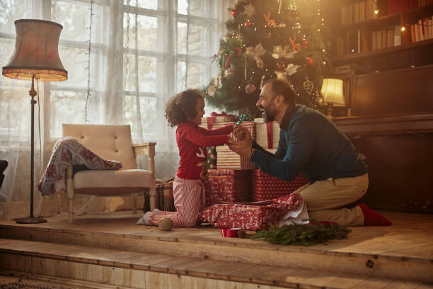 Mixed race father and daughter celebrating winter holidays at home Side view close up of a mixed race daughter with her father sitting in the living room, smiling and celebrating Christmas. Dad is giving gift to cute daughter unwrapping stock pictures, royalty-free photos & images