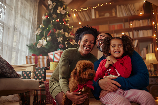 Happy interracial family having fun at home at christmas time, being together, sitting on floor, playing with christmas their cute dog and smiling