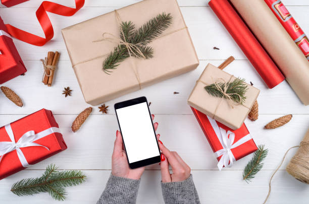 istock Female hands with mobile smart phone with blank screen for app presentation, copy space. Christmas gifts wrapped in kraft and red on wooden background. Flat lay, top view. Application mock up template 1347527698