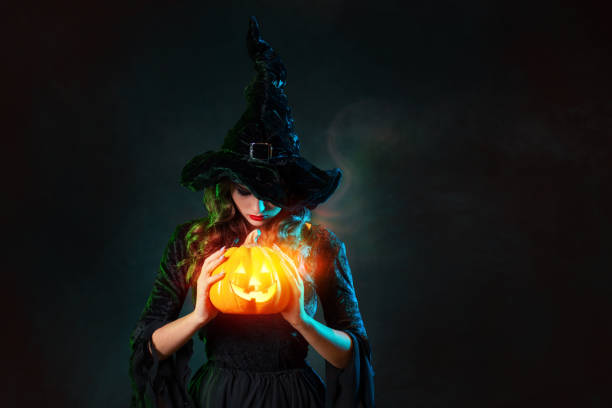 beautiful witch in a pointed hat holds a pumpkin with burning eyes, in her hands