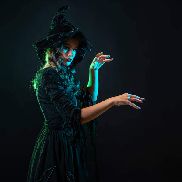 Young woman dressed as a witch sits sadly on a wooden bridge in the forest. She wears a long black dress, black coat, and dark makeup with black lipstick. Next to her are a broom and a witch hat, Halloween atmosphere with autumn colors in the background.
