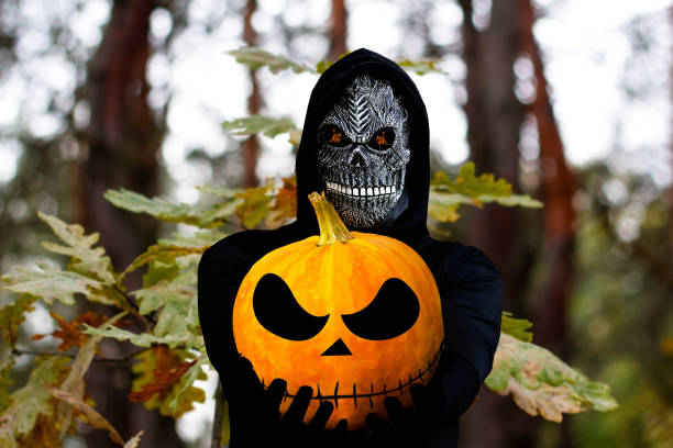 sensenmann mit halloween-kürbiskopf. mann in totenmaske mit feuerflamme in augen auf natur eichenwald blätter hintergrund. halloween feiertagskonzept. dunkler horror - inferno fire flame skull stock-fotos und bilder