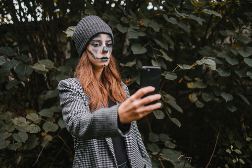 A young woman disguised for Halloween with stage make-up taking selfie in a forest before a Halloween party
