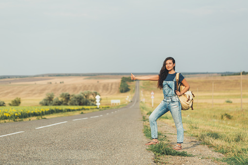Young brunette girl in a denim jumpsuit is hitchhiking around country. Free space for text.
