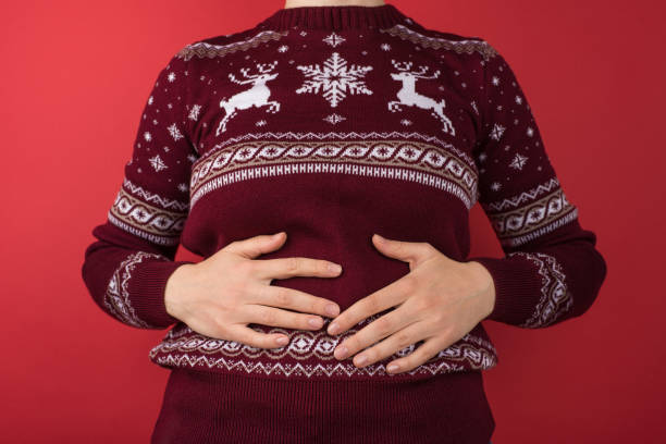 foto ravvicinata ritagliata di una ragazza in maglione natalizio rosso e bianco che tiene il suo stomaco ferito su uno sfondo rosso isolato - eccesso foto e immagini stock