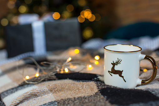 A beautiful white porcelain cup with gilding and with a print in the form of a deer stands on a checkered woolen blanket against the background of a Christmas tree with lights and boxes with gifts