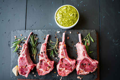 Raw Lamb Chops Prepared on a Dark Slate Cutting Board with a Ramekin or Pesto on the side with Rosemary and other herbs