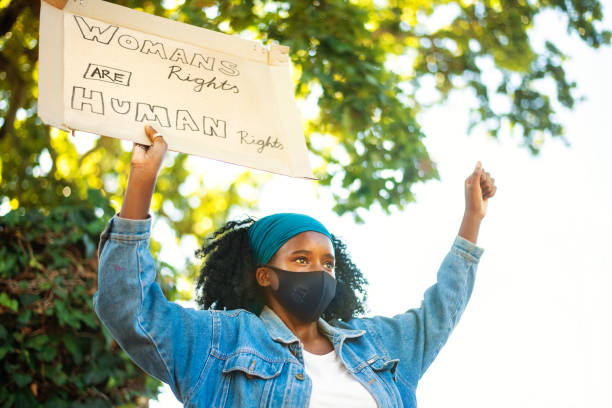 Teenage girl in a face mask holding up a women's rights protest sign Young African teenage girl wearing a protective face make holding up a women's rights protest sign while standing outside me too social movement stock pictures, royalty-free photos & images