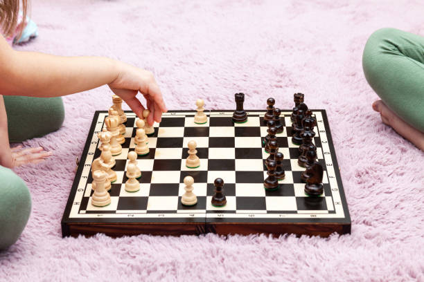 deux enfants anonymes d’âge scolaire méconnaissables, des petites filles jouant aux échecs dans leur chambre à la maison, un échiquier allongé sur le tapis sur le sol, un enfant faisant un mouvement, déplaçant le pion - chess board room business strategy photos et images de collection