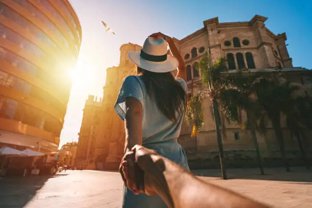 Follow me - POV. Young tourist woman in white sun hat holding her boyfriend by hand and walking in Malaga city at sunset. Couple on summer holiday vacation in Spain. Traveling together. High quality