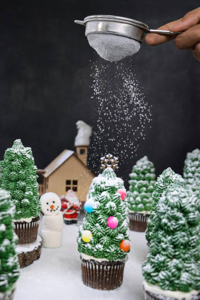 istock Image of unrecognisable person sifting icing sugar snow over night conifer forest scene display of batch homemade, Christmas tree design chocolate cupcakes in paper cake cases, model house with Santa Claus and marshmallow snowman 1347573868