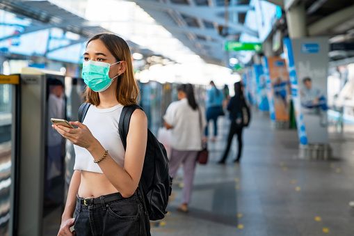 Young Asian woman wearing surgical face mask during COVID-19 pandemic protection waiting for subway or skytrain at railway station platform with using smartphone for online shopping or social media. City life and technology concept.