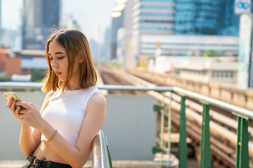 Young Asian woman waiting for skytrain at railway station platform using smartphone with internet for online shopping or social media. Modern female enjoy outdoor lifestyle and shopping in the city
