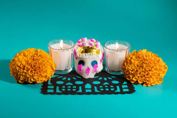 Photo of Day of the dead pottery skull (calavera) votive candles and marigold cempasuchil flowers on black papel picado on teal blue background, Dia de Muertos concept