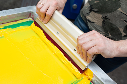 Serigraphy silk screen print process at clothes factory. Frame, squeegee and plastisol color paints.