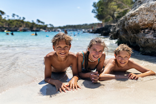 Brothers and sister are having fun in the sea. Kids are lying on front on the sand in the beautiful cove on the shores of Majorca, Spain\nSunny summer day.\nCanon R5
