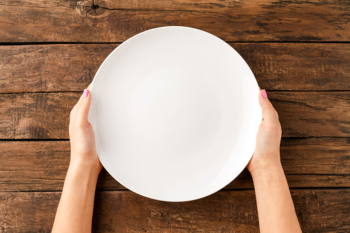 Woman’s hands holding empty white plate over rustic wooden table. Top view