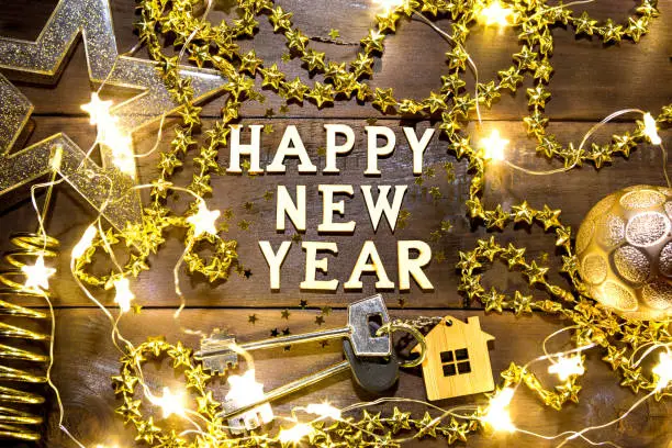 House key with keychain cottage on a festive background with sequins, stars, lights of garlands. Happy New Year-wooden letters, greetings, greeting card. Purchase, construction, relocation, mortgage