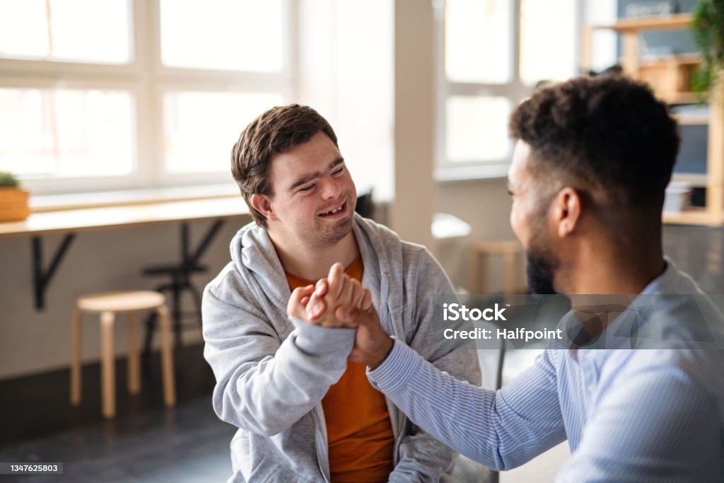 Young happy man with Down syndrome with his mentoring friend celebrating success indoors at school. A young happy man with Down syndrome with his mentoring friend celebrating success indoors at school. Disability Stock Photo