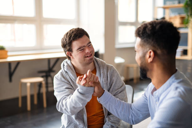 Young happy man with Down syndrome with his mentoring friend celebrating success indoors at school. stock photo