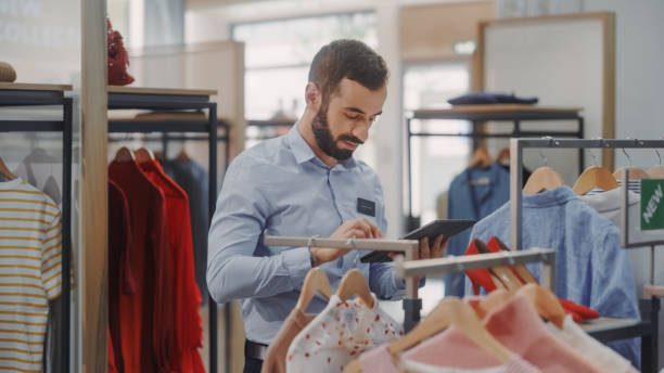 istock Clothing Store: Male Visual Merchandising Professional Uses Tablet Computer To Create Collection. Fashionable Shop Sales Retail Manager Checks Stock. Big Mall SUpermarket with Stylish Merchandise 1347626132