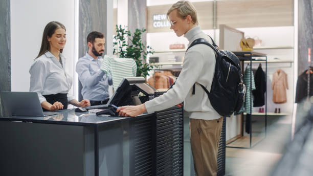 istock Clothing Store: Handsome Young Man At Checkout Counter Buys Clothes from Friendly Retail Sales Assistant, Paying with Contactless NFC Smartphone Touching Terminal. Shopping Mall with of Designer Brand 1347626146