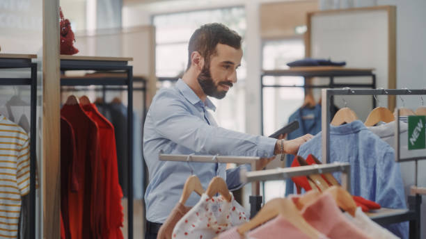 istock Clothing Store: Male Visual Merchandising Professional Uses Tablet Computer To Create Collection. Fashionable Shop Sales Retail Manager Checks Stock. Big Mall Supermarket with Stylish Merchandise 1347626150
