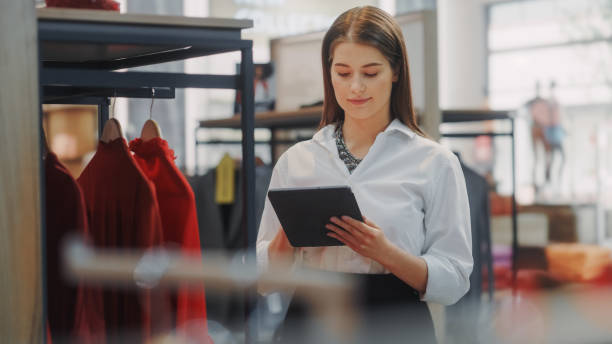 istock Clothing Store: Female Visual Merchandising Specialist Uses Tablet Computer To Create Stylish Collection. Fashion Shop Sales Retail Manager Checks Stock. Small Business Owner Orders Merchandise 1347626151