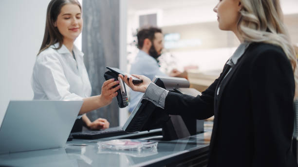 clothing store: young woman at counter buys clothes from friendly retail sales assistant, paying with contactless nfc smartphone touching terminal. trendy fashion shop with of designer brands. - clothing store paying cashier credit card imagens e fotografias de stock