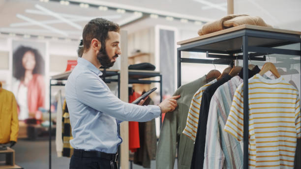 istock Modern Clothing Store: Male Visual Merchandising Professional Uses Digital Tablet Computer To Create New Collection Strategy. Fashionable Shop Sales Retail Associate Orders Stylish New Stock Items 1347626168