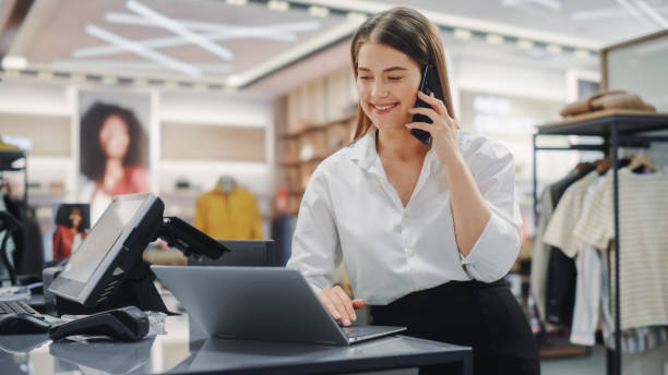 istock Modern Clothing Store: Friendly Customer Support Specialist Talks to Client on the Smartphone. Smiling Retail Sales Manager or Young Designer Contacts Client about Online Order in Fashion Shop 1347626189