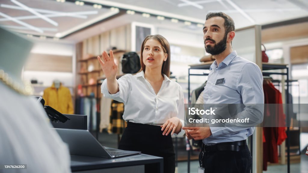 Clothing Store: Businesswoman Uses Laptop Computer, Talks to Visual Merchandising Specialist, Collaborate To Create Stylish Collection. Business Owner's Fashion Shop: Sales Manager Talks to Designer Retail Stock Photo