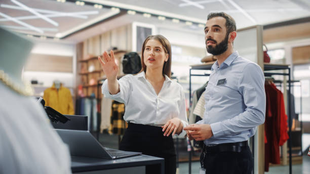 clothing store: businesswoman uses laptop computer, talks to visual merchandising specialist, collaborate to create stylish collection. business owner's fashion shop: sales manager talks to designer - owner boutique store retail imagens e fotografias de stock