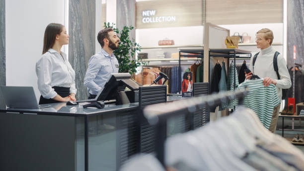 istock Clothing Store: Handsome Young Man At Checkout Counter Buys Clothes from Friendly Retail Sales Assistant. Shopping Mall with of Designer Brand. Trendy Fashion Shop with of Designer Brands. 1347626215