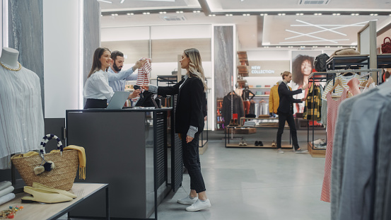 Mostrador de caja de la tienda de ropa: Hermosa joven compra blusa de un amable asistente de ventas minoristas, pagando con tarjeta de crédito sin contacto. Tienda de moda con marcas de diseñadores. photo