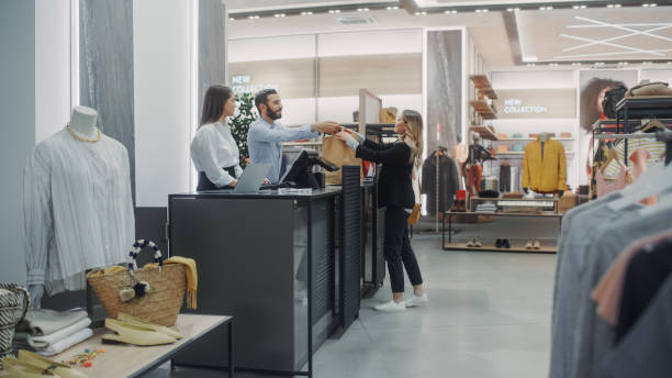 clothing store checkout cashier counter: beautiful young woman buys blouse from friendly retail sales assistant, receiving bag with purchases. trendy fashion shop with of designer brands - clothing store paying cashier credit card imagens e fotografias de stock