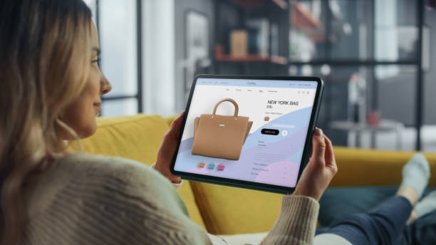 istock Beautiful Caucasian Female is Using Tablet Computer with Clothing Online Web Store to Choose and Buy Coat from New Collection. Female Surfing the Net and Lying on Couch Sofa at Home Living Room. 1347626307