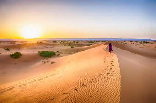 Women walking on sand dunes in arid desert at sunrise and wearing red dress, scenic landscape of Sahara desert in Morocco