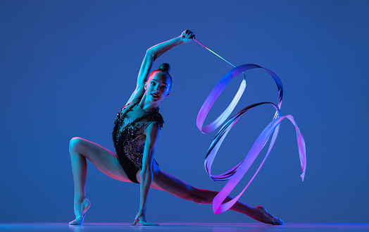 Exercise with colored ribbon. Studio shot of little girl, rhythmic gymnastics artist isolated on blue studio background in pink neon light. Concept of sport, action, aspiration, wellness, fitness.